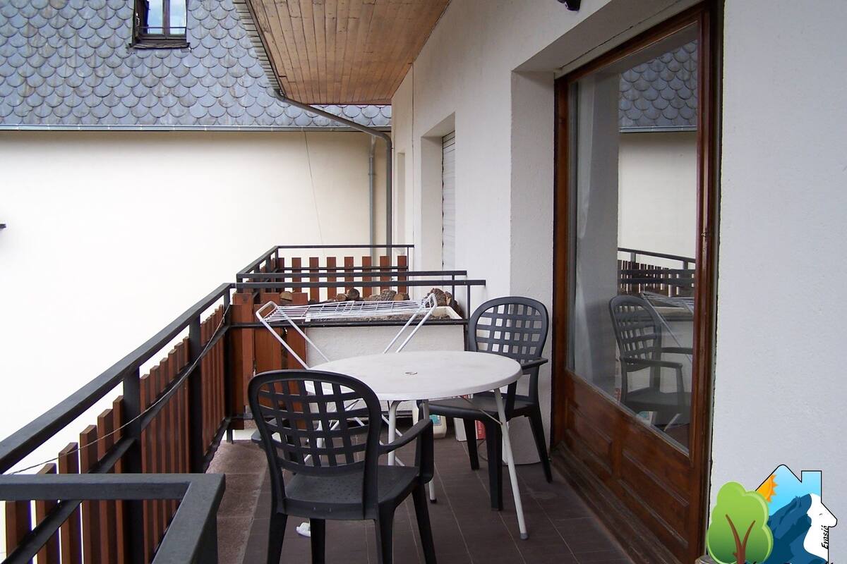Un balcón con mesa y sillas, baranda de madera y vista a un edificio con tejado de baldosas.