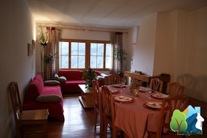 Un comedor con una mesa de madera preparada para cuatro personas, rodeada de sillas de madera. Se ve un sofá rojo y una silla de madera en primer plano.