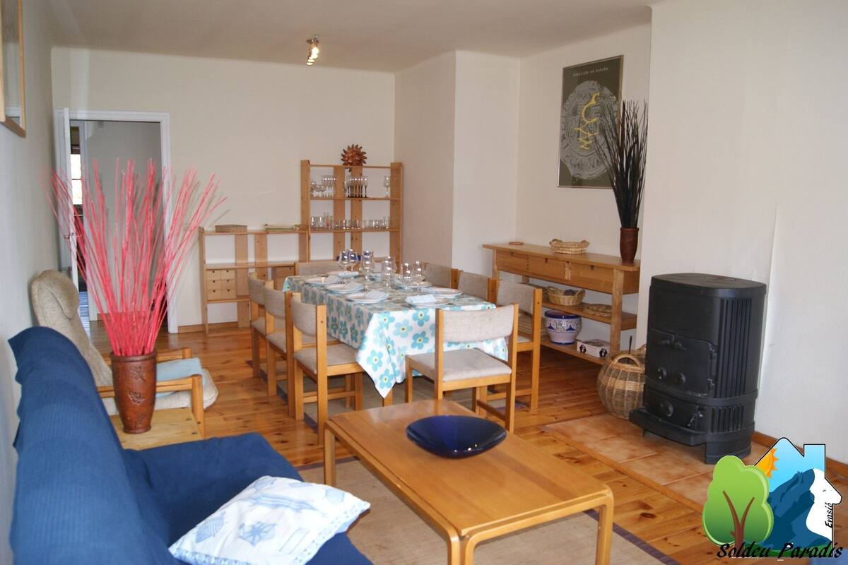 Una sala de estar acogedora con un sofá azul, una mesa de comedor de madera dispuesta para cuatro comensales, un estufa de leña negra y una zona de comedor con un mueble y estanterías de madera.