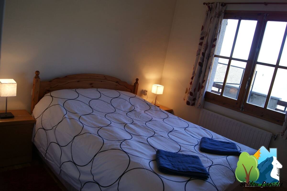 Un dormitorio con cama, dos lámparas de noche, una ventana con cortinas y vistas a edificios.