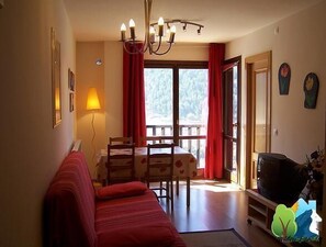 Una sala de estar acogedora con un sofá rojo, una mesa de comedor con sillas, un televisor y un balcón con cortinas rojas.