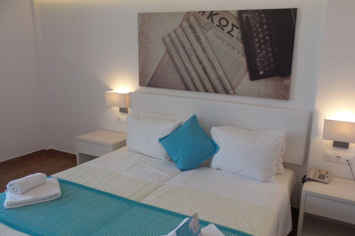 A neatly made bed with white and blue bedding, two bedside lamps, a wall-mounted telephone, and a folded towel on the floor.