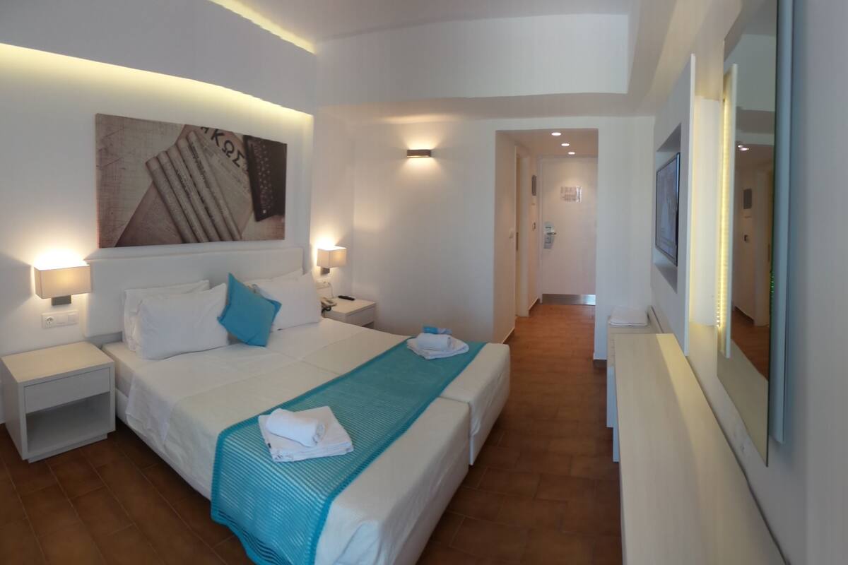 A hotel room with a large bed, bedside tables, a wall-mounted TV, and a hallway leading to another room.