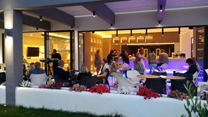 An outdoor seating area with tables and chairs, a stage with a keyboard, and a bar area with people. - Cosmopolitan Kos Hotel (Kos)