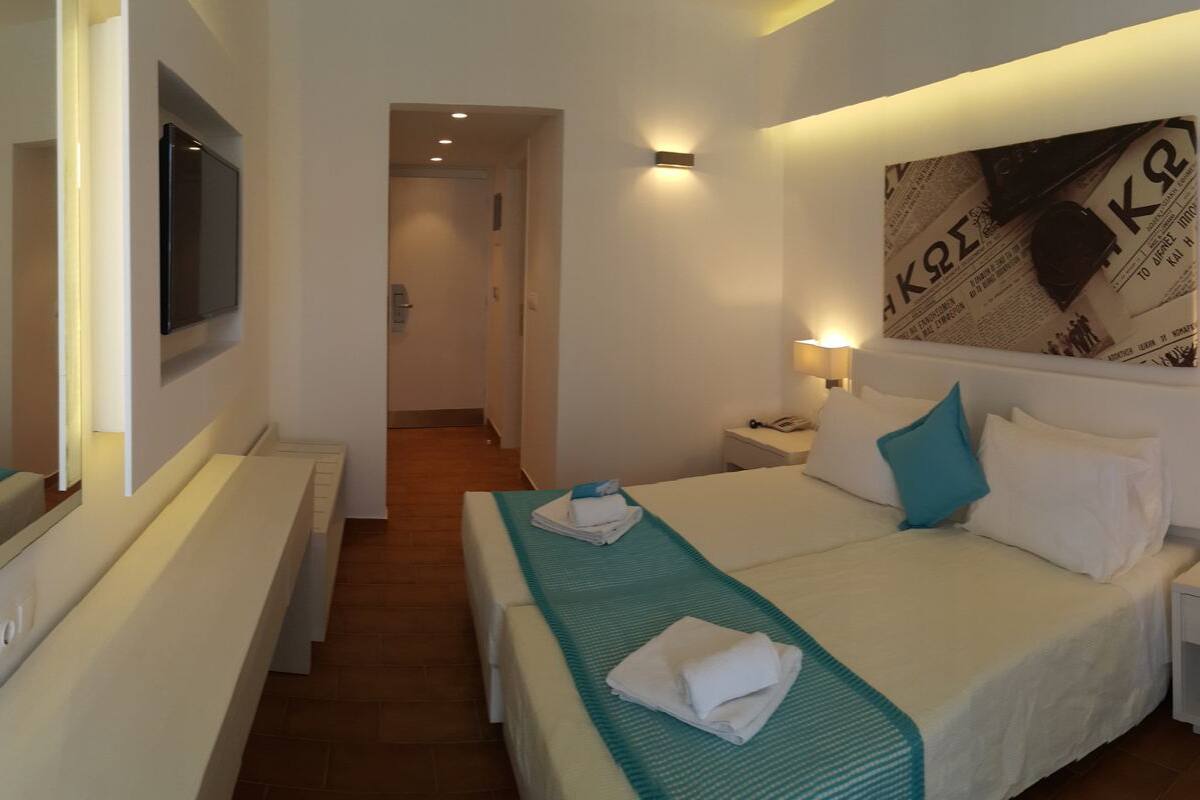 A hotel room with a bed, a bedside table, a nightstand, a wall-mounted TV, and a door leading to another room.