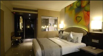A hotel room with a large bed, a bedside table, a nightstand, a mirror, and a colorful abstract painting on the wall.