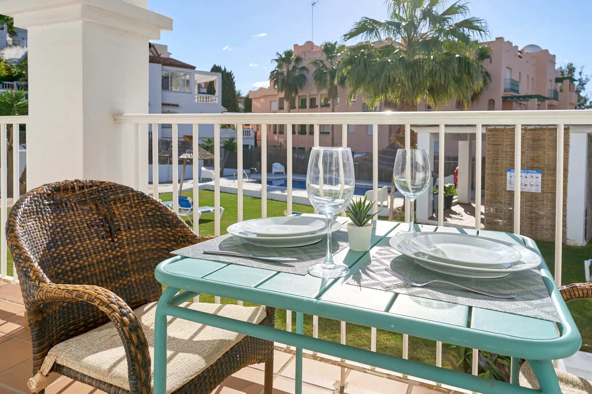 Un balcón con sillas de mimbre, una mesa con platos y vasos, y vistas a una piscina y edificios.