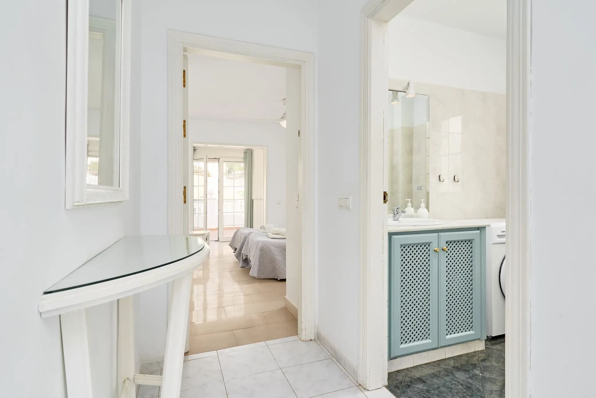 Un couloir d’hôtel avec une table en verre, une salle de bain avec un lavabo et un miroir, et un lit à l’arrière-plan.