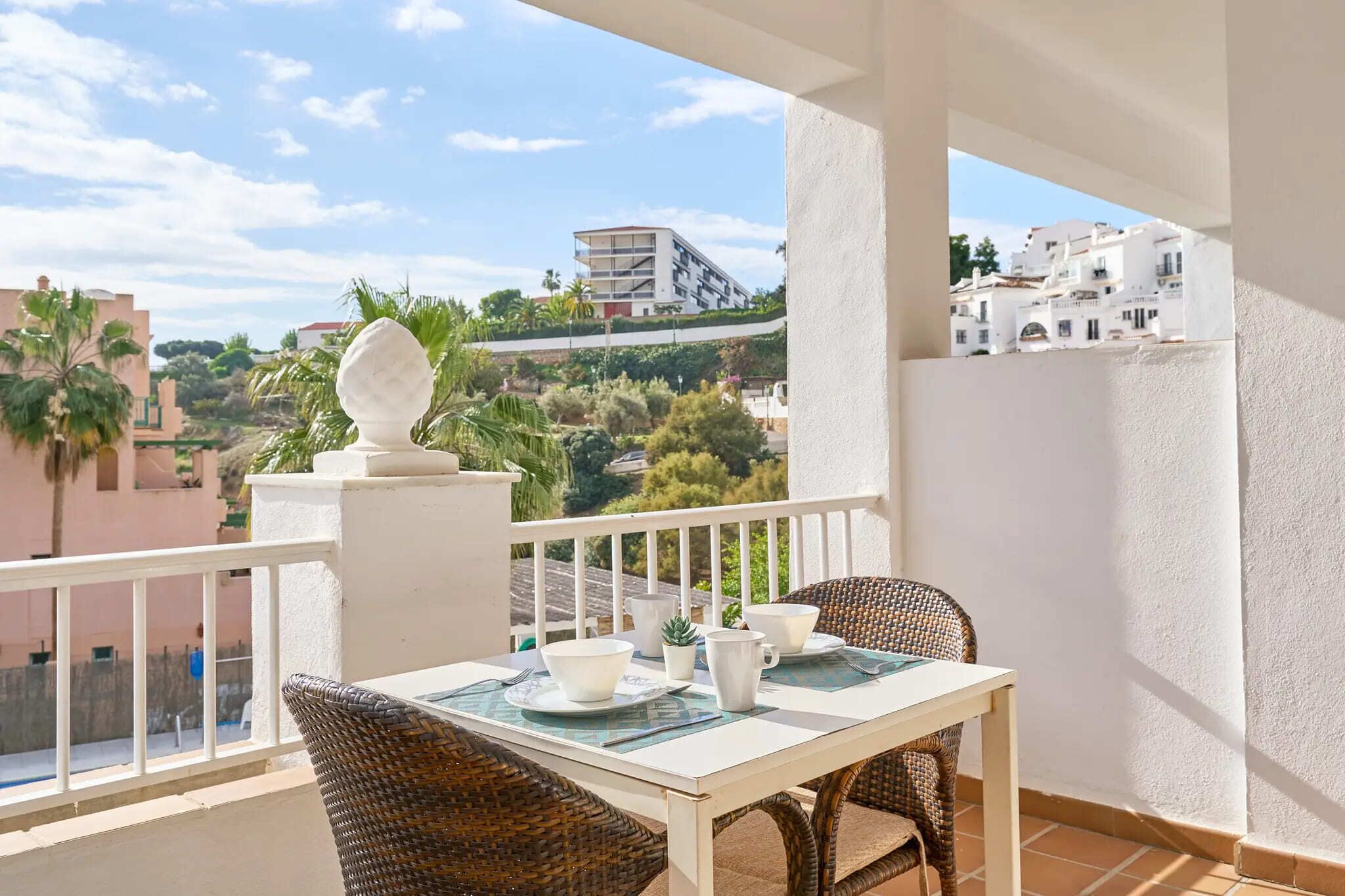 Un balcón con una mesa para dos, sillas de mimbre y vistas a edificios y zonas verdes.