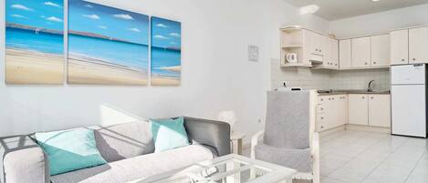 A modern living room with a sofa, armchair, and coffee table, a kitchen with white cabinets and appliances, and a large beach-themed painting on the wall.