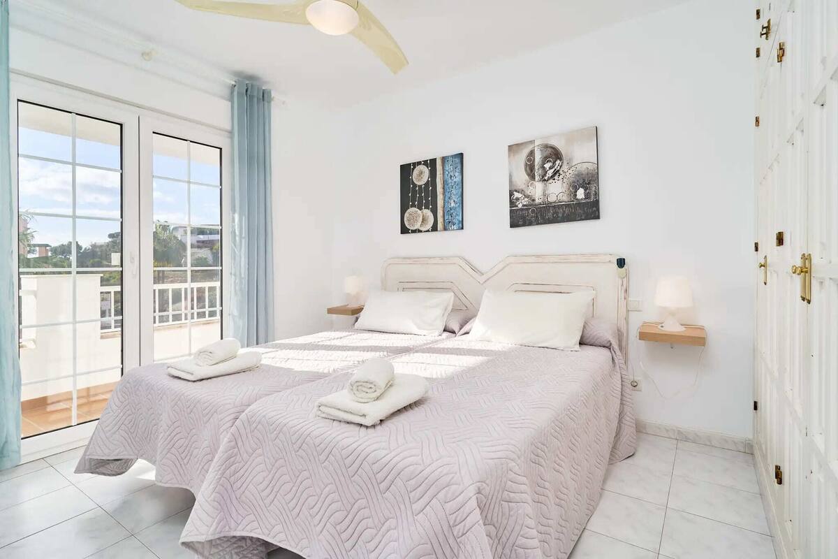 A bedroom with a large bed, two bedside tables, a ceiling fan, and a view through a sliding glass door.