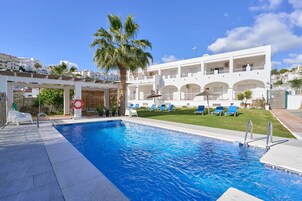 Una piscina con sillas de descanso, una palmera y un edificio blanco con balcones.