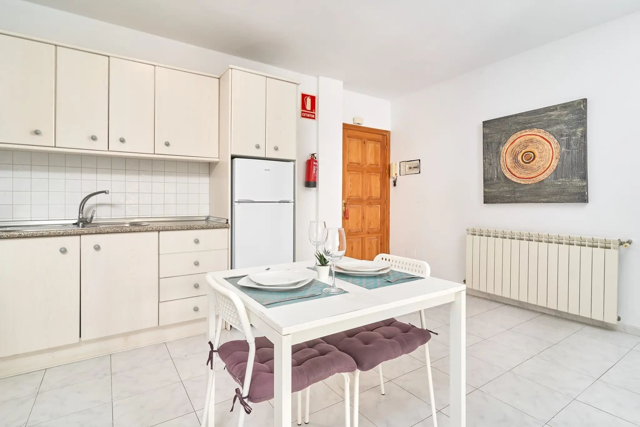 Une cuisine de taille réduite avec des meubles blancs, un réfrigérateur blanc, une table à manger avec des coussins violets et une porte en bois.