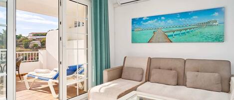 A living room with a sofa, a glass coffee table, a painting of a pier, and a sliding glass door leading to a balcony with a chair and a sunbed.