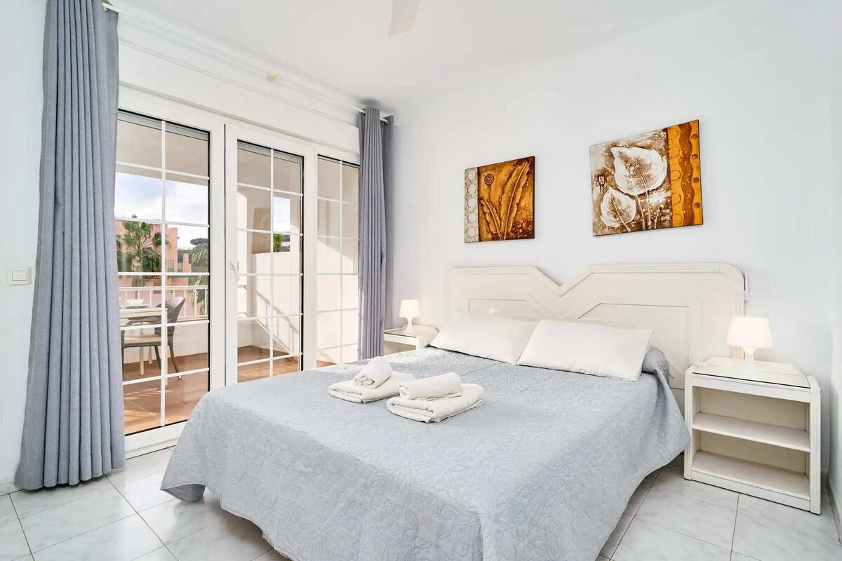A bedroom with a bed, two wall art pieces, a nightstand with a lamp, and a view through a sliding glass door.