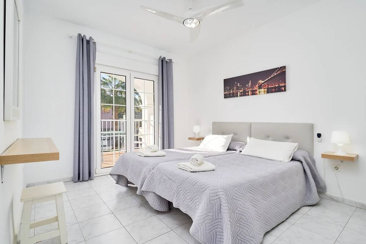 A bedroom with a bed, two bedside tables, a ceiling fan, a window with curtains, and a cityscape painting.