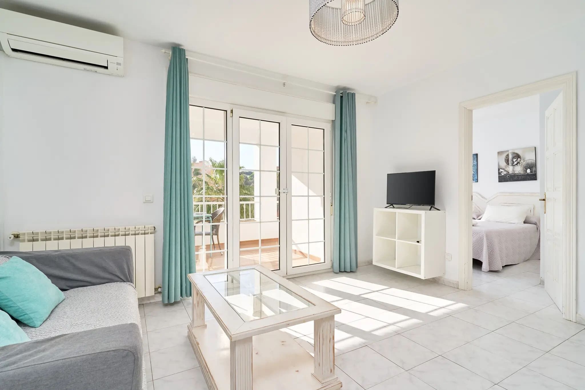 A living room with a sofa, a glass-top coffee table, a TV on a white stand, and a sliding glass door leading to an outdoor area.