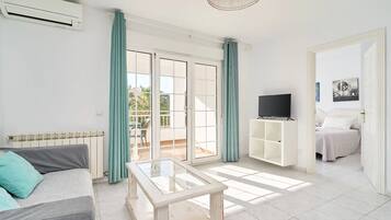 A living room with a sofa, a glass-top coffee table, a TV on a white stand, and a sliding glass door leading to an outdoor area.