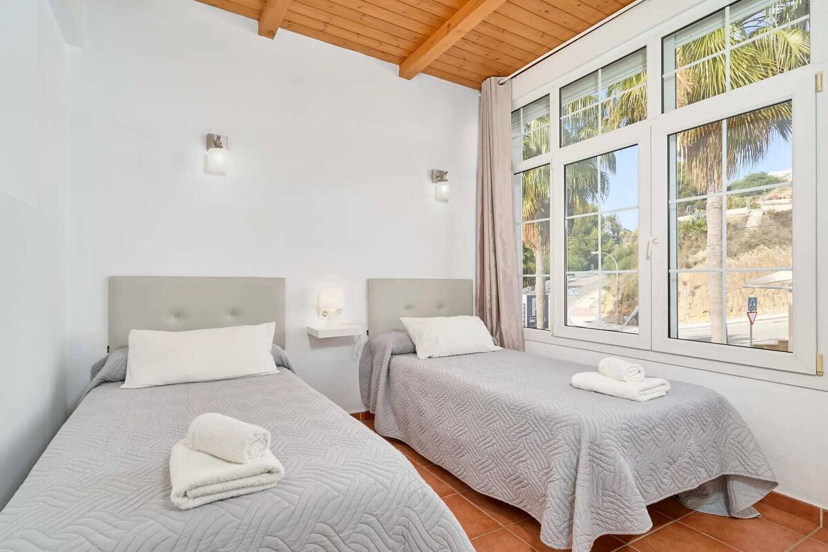 A room with two beds, each with a headboard, a nightstand, and a lamp. There is a large window with a view of palm trees and a building.