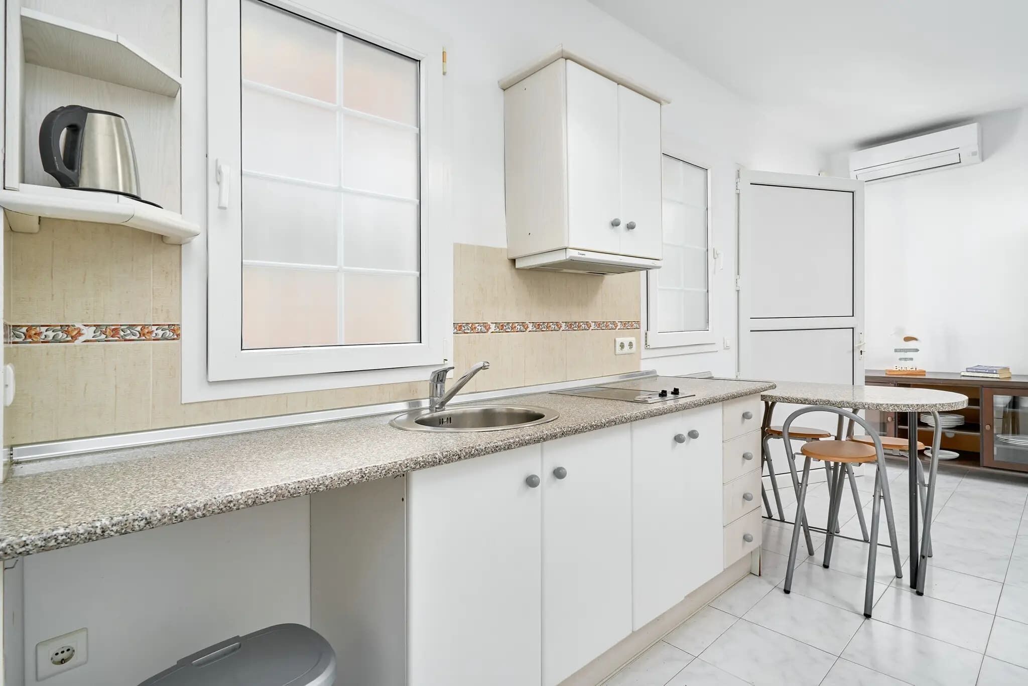 A kitchen with white cabinets, a stainless steel sink, a window, and a small dining area with stools.