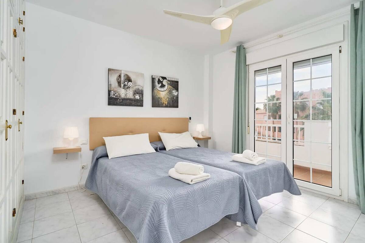 A hotel room with two beds, a ceiling fan, artwork on the wall, and a balcony with curtains.