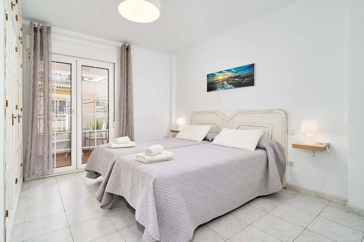 A bedroom with a bed, two bedside tables, a nightstand, a painting, and a sliding glass door leading to a balcony.