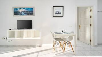 Una habitación luminosa con una televisión en un estante, una mesa de comedor con sillas y dos cuadros en la pared.