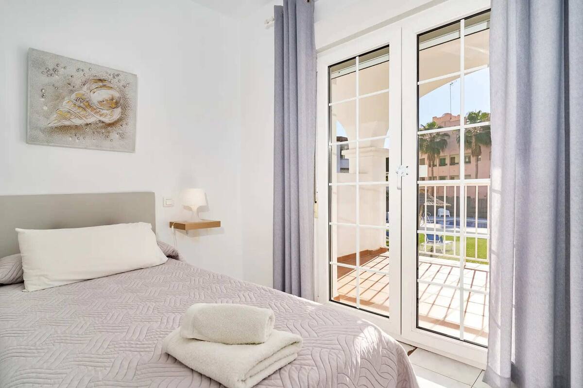 A hotel room with a bed, a painting on the wall, a window with curtains, and a view of the outside.
