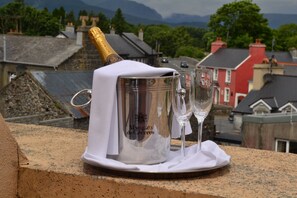 A champagne bottle and two glasses on a table with a scenic view of houses and mountains in the background. - Fairhill House Hotel (Clonbur)