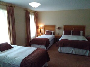 Ein Hotelzimmer mit drei Einzelbetten, einem Holz-Kopfteil, einem Nachttisch mit Lampe und einem Fenster mit Vorhängen.