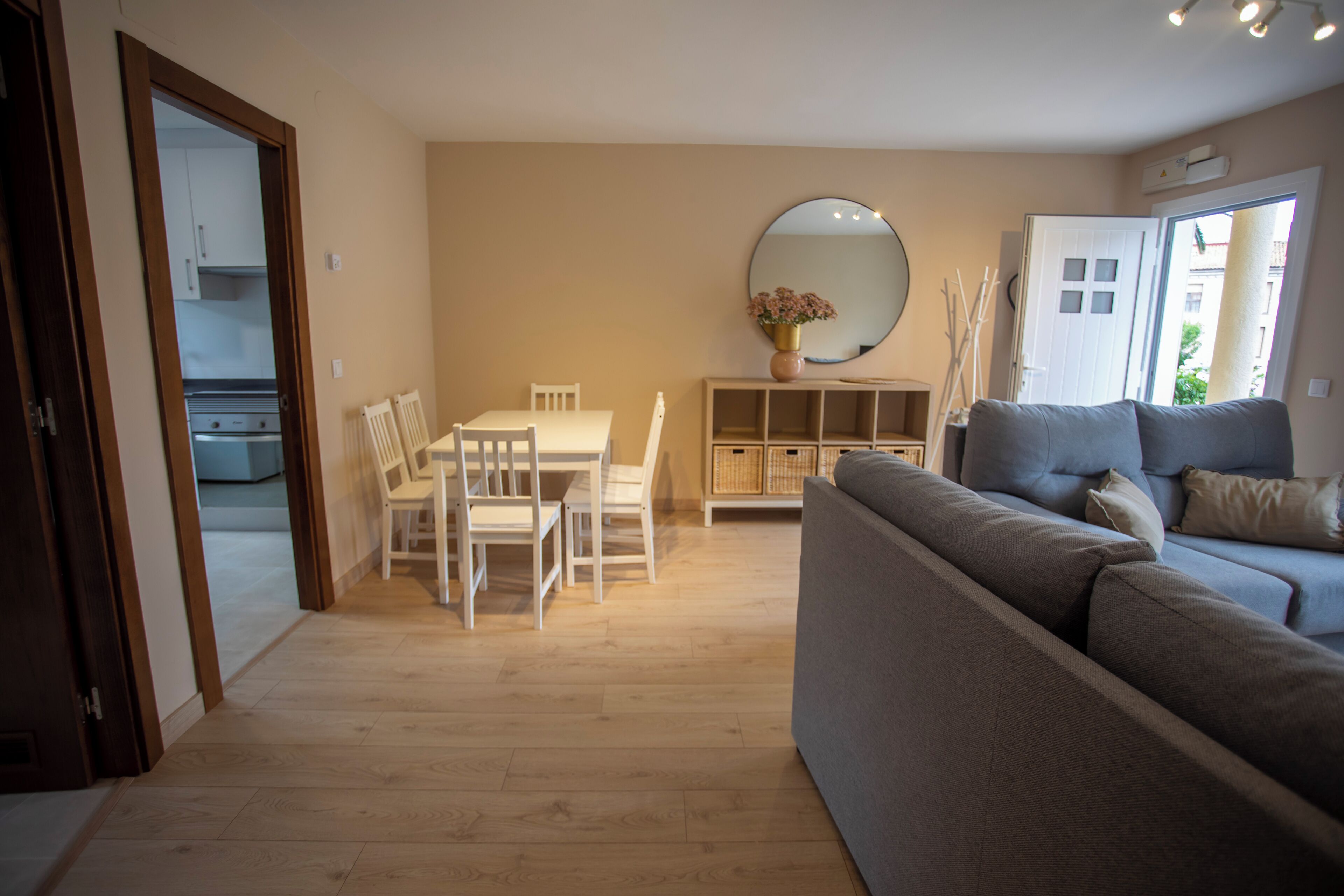 Een woonkamer met een bank, een eettafel met stoelen, een spiegel, en een keuken zichtbaar door een geopende deur.