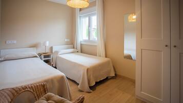 A hotel room with two beds, a nightstand, a wicker chair, a window with curtains, and a door.