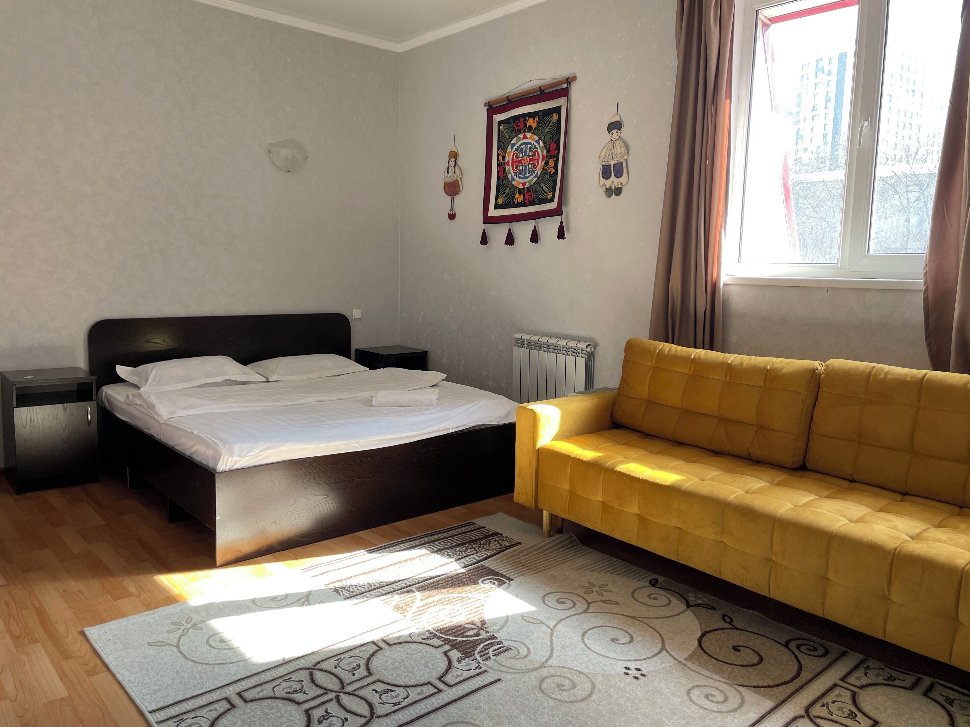 A bedroom with a bed, a yellow sofa, a radiator, a window with curtains, and decorative wall hangings.