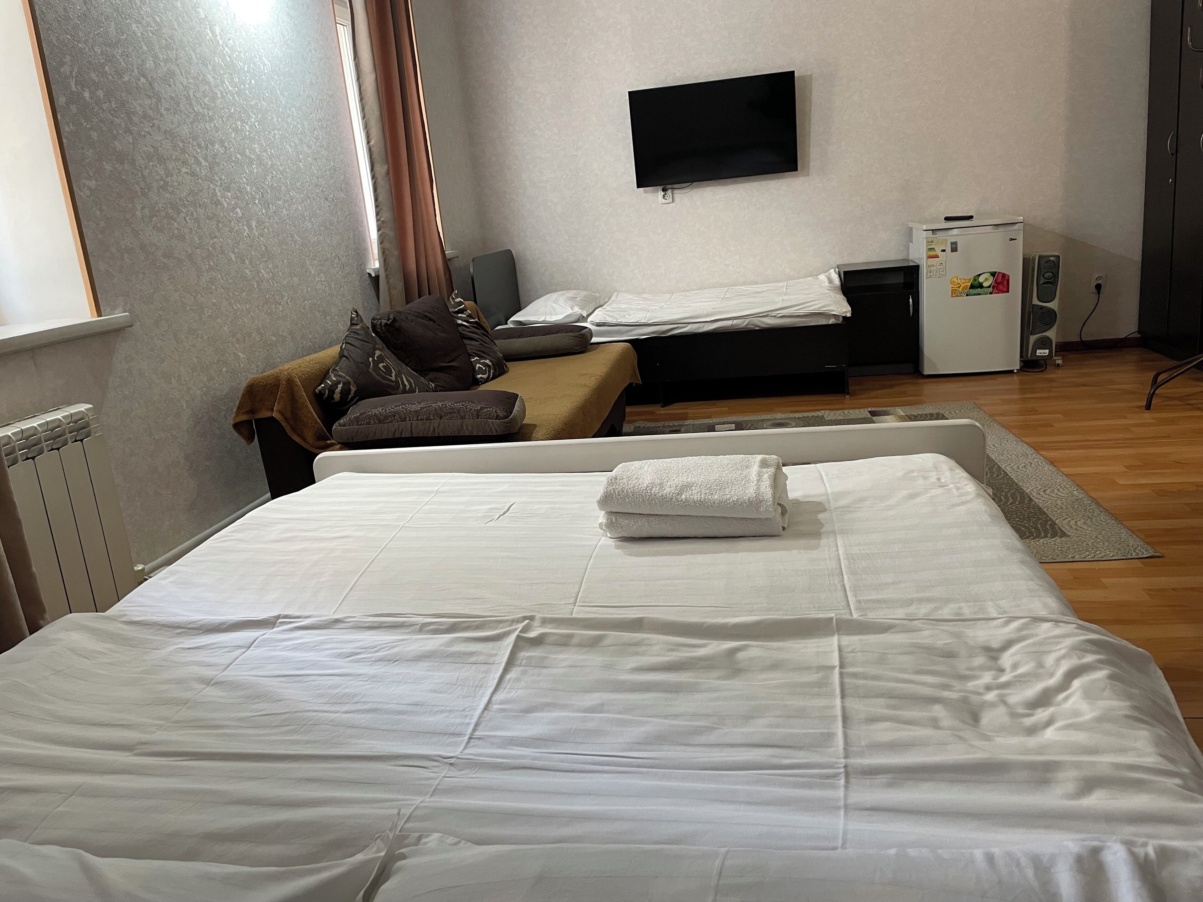 A hotel room with a large bed, a small sofa, a TV mounted on the wall, and a refrigerator.