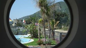 Vista a través de una ventana que muestra una zona residencial con piscina, palmeras y una montaña al fondo.