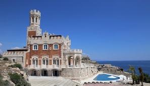 Un edificio histórico con una torre, una piscina y vista al mar.