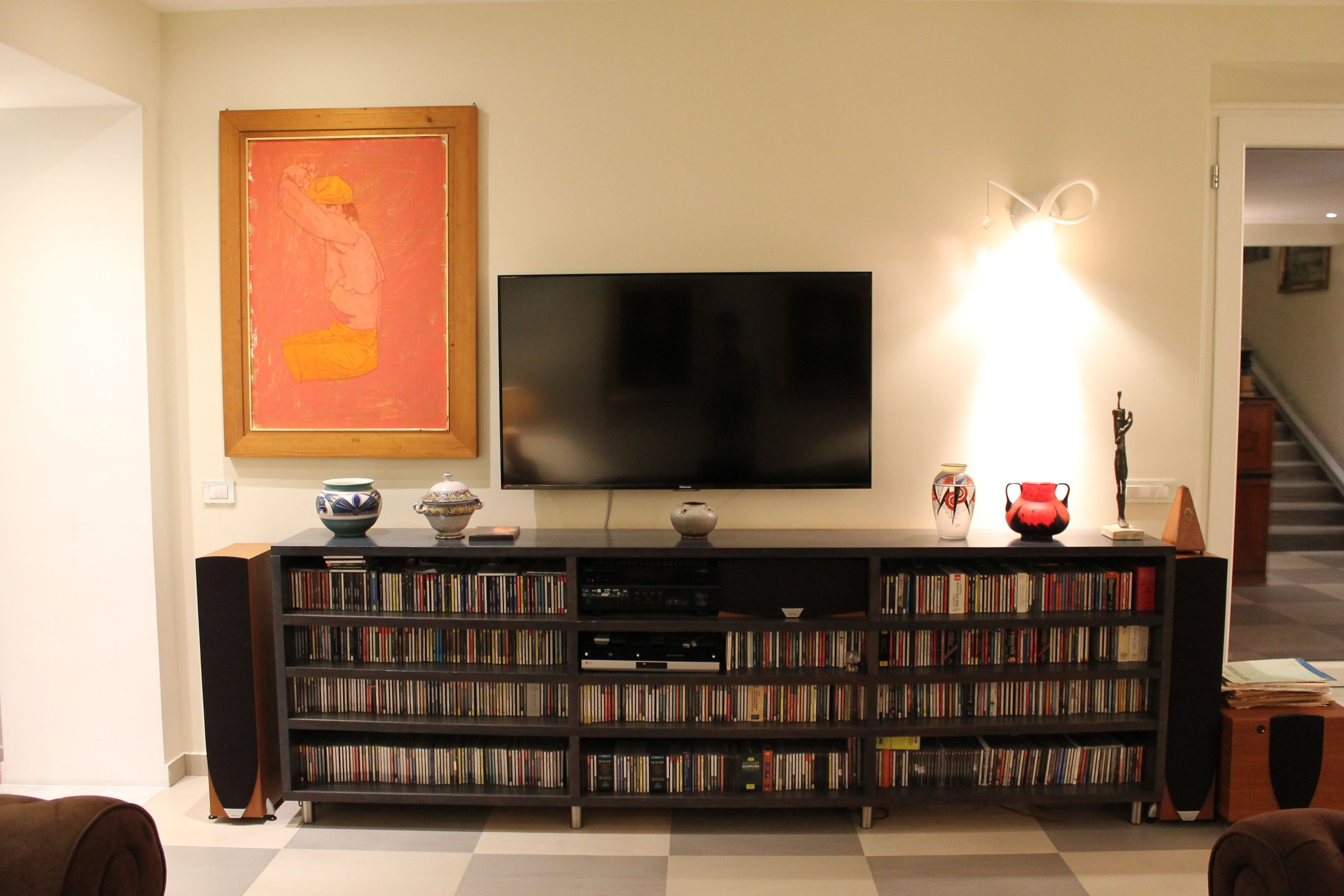 Una sala de estar con un televisor de pantalla plana grande montado en la pared, una estantería llena de libros y DVD, y un jarrón decorativo en el estante.