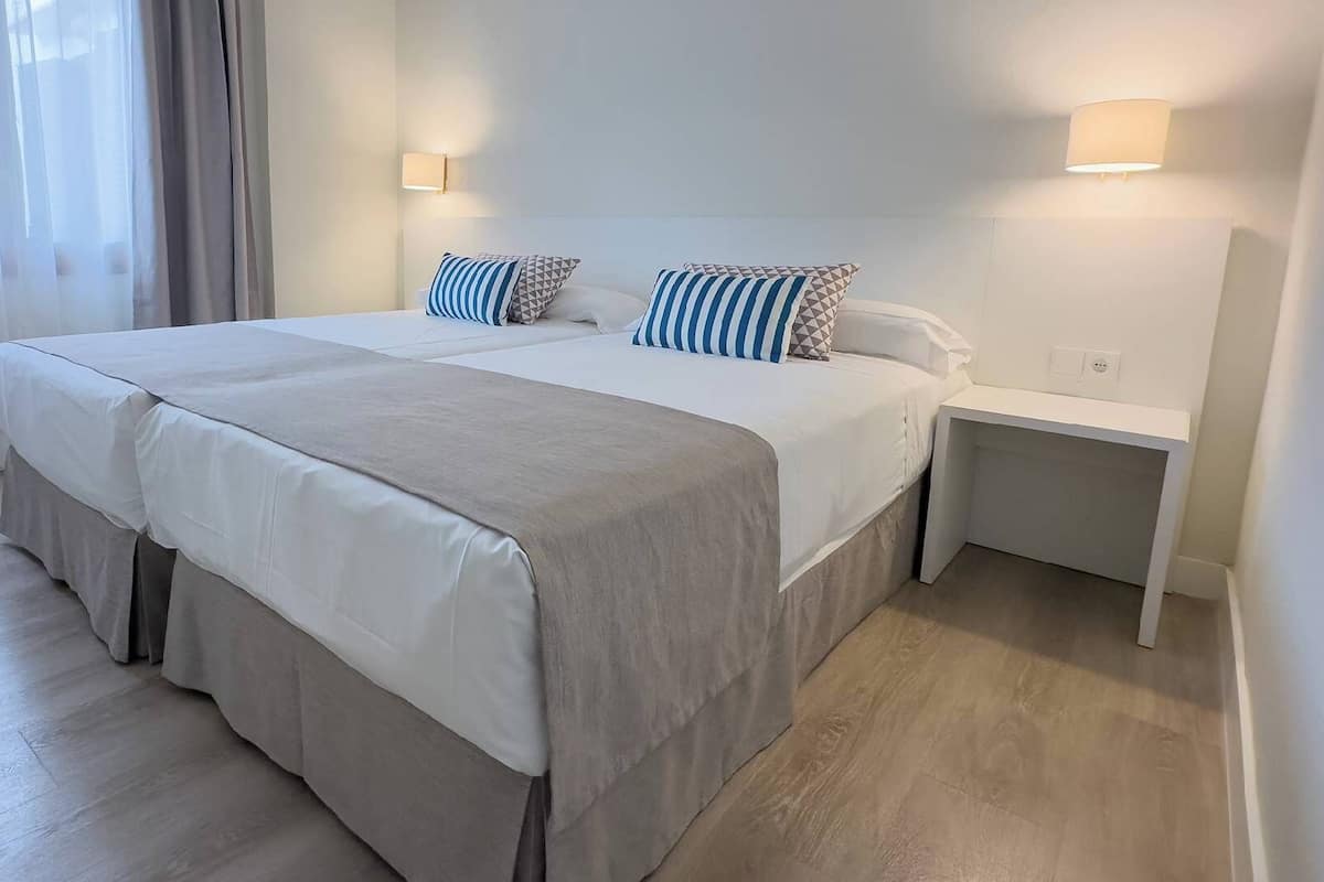 Ein Doppelbett mit einer schlichten Bettdecke und zwei Kissen, ein Nachttisch und eine Wandlampe.