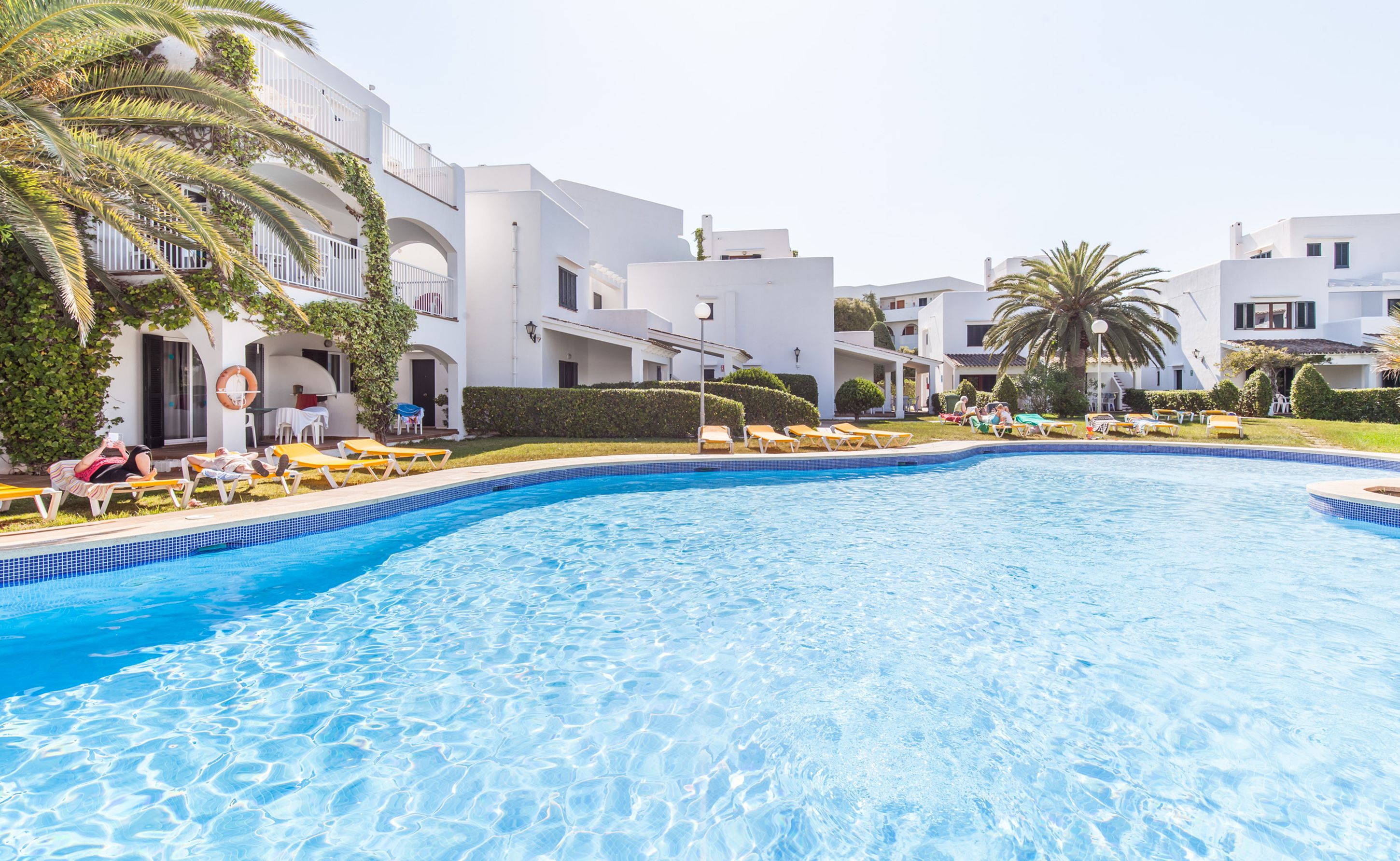 Una piscina rodeada de edificios blancos con balcones, palmeras y tumbonas.