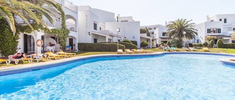Una piscina rodeada de edificios blancos con balcones, palmeras y tumbonas.