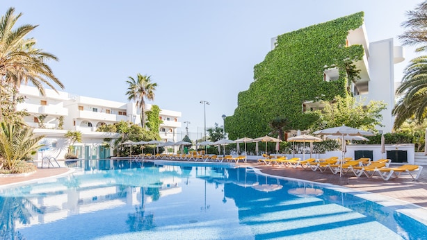 Una piscina con lettini e ombrelloni, circondata da palme e un edificio con facciata verde.