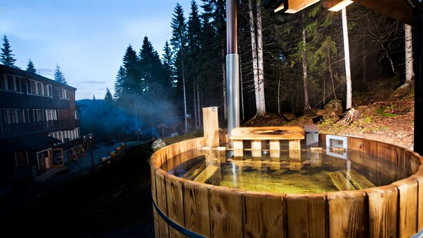 A wooden hot tub surrounded by a forest, with a building in the background. - Eko sport hotel Björnson (Demanovska Dolina)