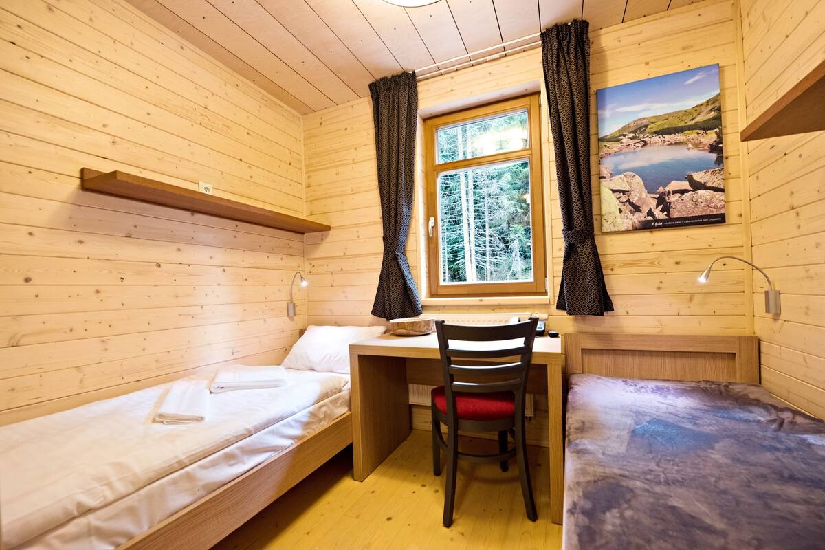 Una habitación de cabaña de madera con dos camas, un escritorio, una silla y una ventana con cortinas.