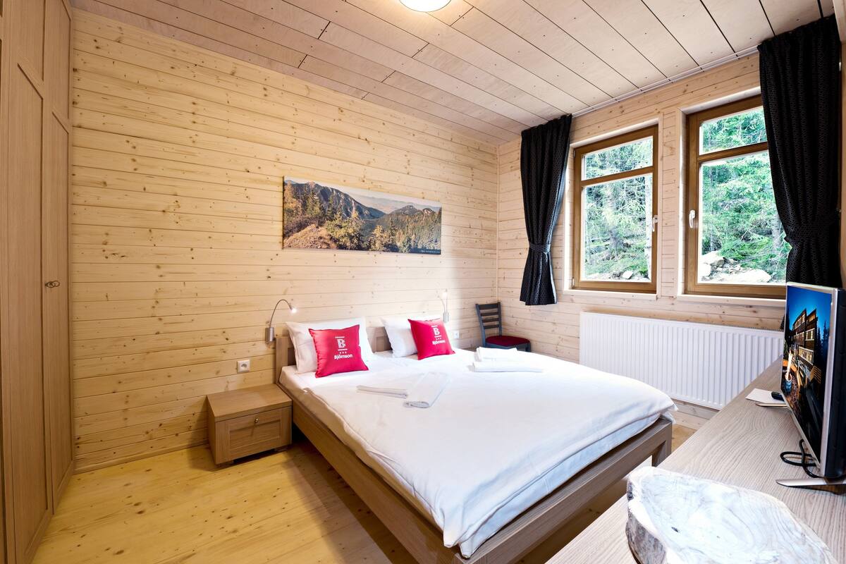 Un dormitorio de cabaña de madera con cama, almohadas, una mesita de noche, una pequeña mesa, una silla, una ventana con cortinas y un cuadro en la pared.