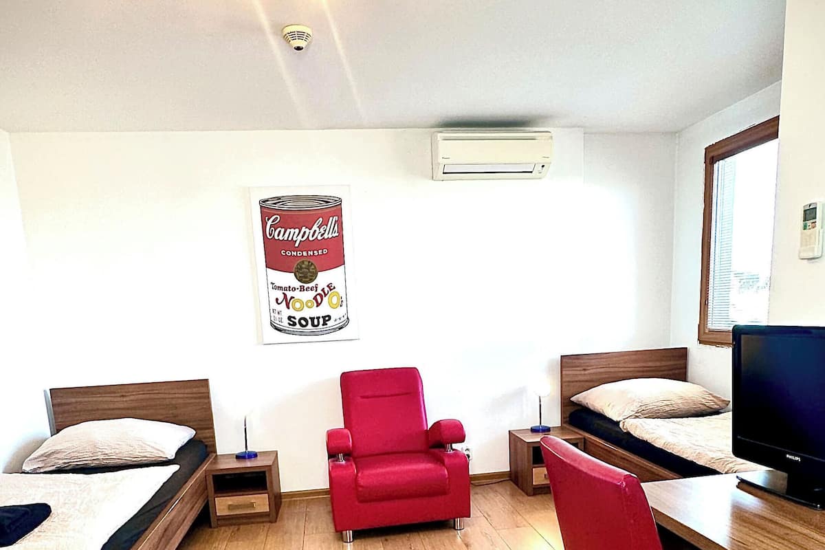 A room with two single beds, a red armchair, a desk with a computer, and a poster on the wall.