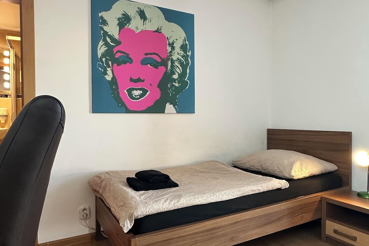 A bedroom with a bed, a chair, a nightstand, and a wall-mounted artwork.