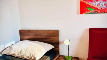A bedroom with a wooden bed, a bedside table with a lamp, a red chair, and a colorful painting on the wall.