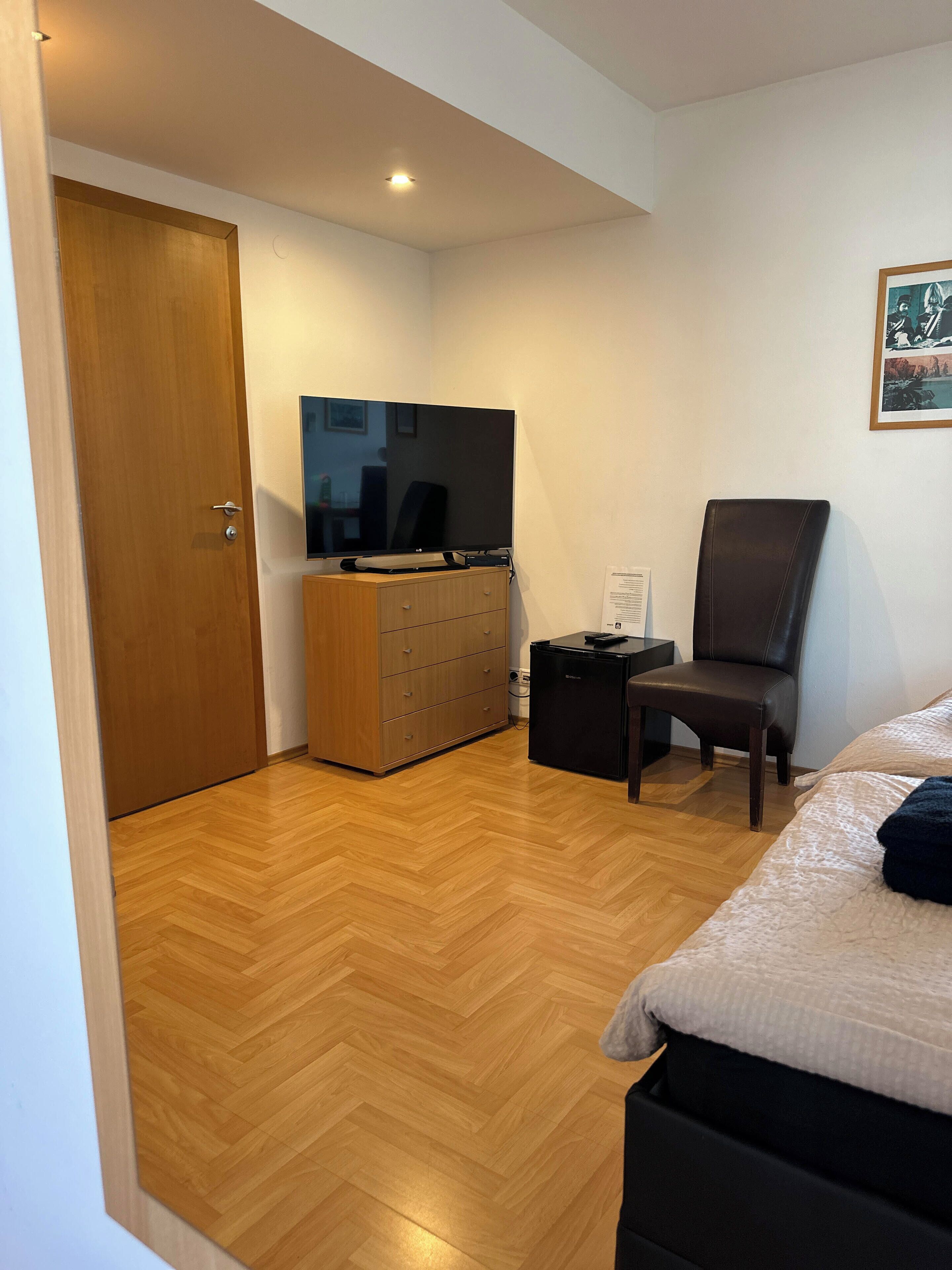 Ein Schlafzimmer mit Holzboden, einem Bett, einem Schreibtisch mit Stuhl, einem Fernseher und einem Kleiderschrank.