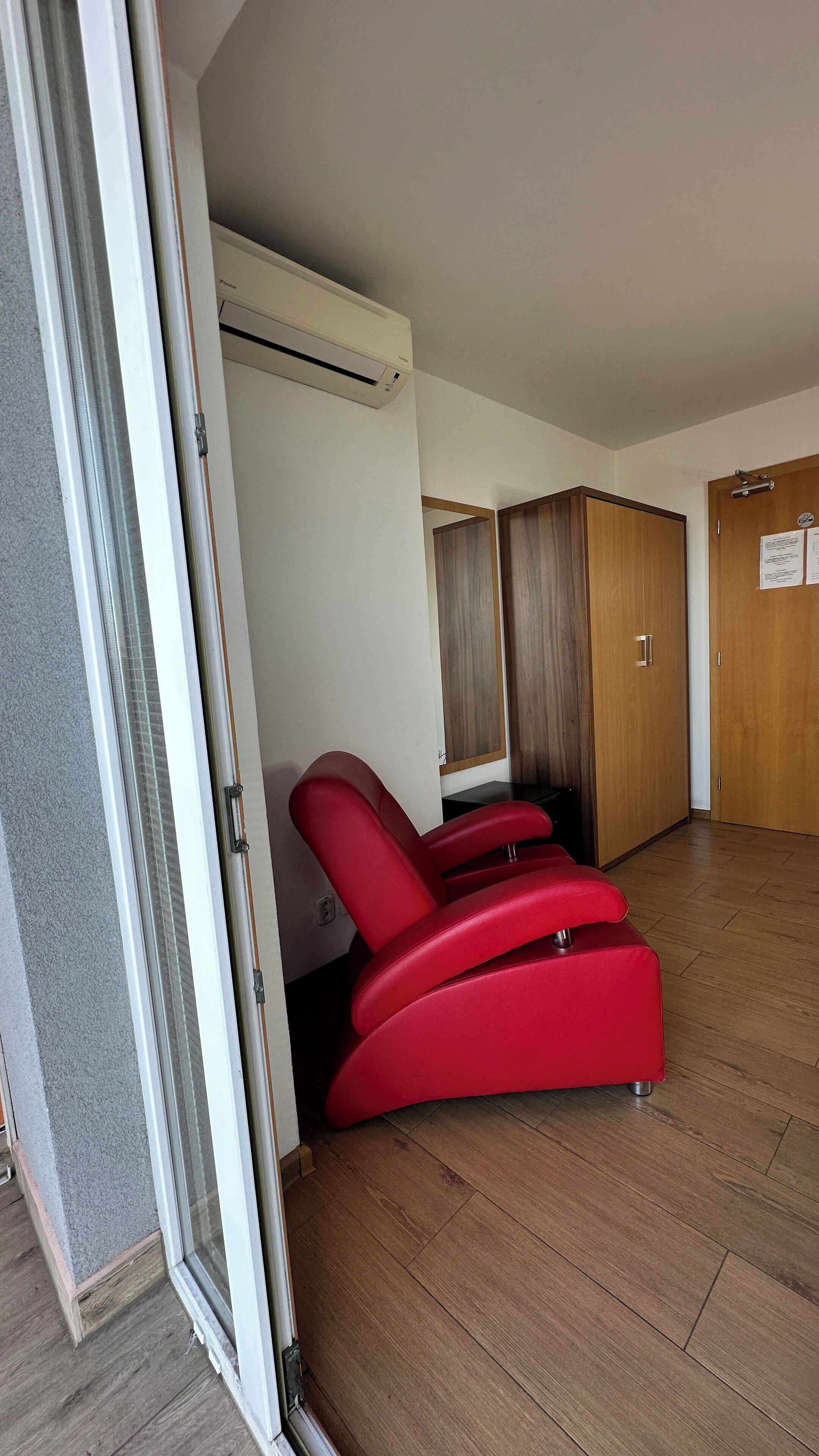 A room with a sliding glass door, a red reclining chair, a wooden wardrobe, and a white air conditioning unit.
