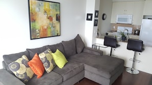 A modern living room with a grey sectional sofa, a small kitchen area with a microwave, and a dining table with chairs. - Custom Luxury Suite - By Elite Suites (Mississauga)